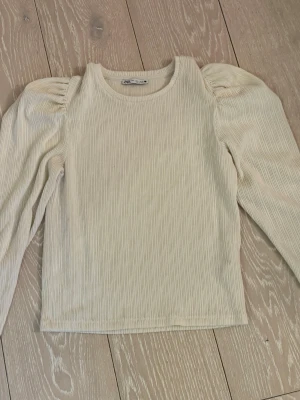 Beige ribbad topp med puffärm Zara - Trendig beige ribbad topp från Zara med långa puffärmar och rund halsringning. Toppen har en figurnära passform och är tillverkad i ett mjukt material som känns skönt mot huden. Perfekt för dig som gillar stilrena och moderna plagg.