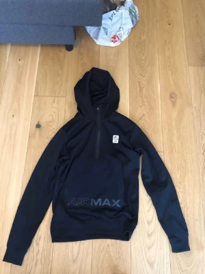 Svart Nike Air Max hoodie med dragkedja - Svart hoodie från Nike Air Max med halv dragkedja framtill och huva. Stort AIR MAX-tryck på magen och liten patch på bröstet. Skön passform och perfekt för en sportig look. Tillverkad i mjukt material som känns bekvämt hela dagen.