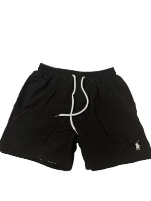 Svarta badshorts från Polo Ralph Lauren - Svarta badshorts från Polo Ralph Lauren med vit snörning i midjan och klassisk logga broderad på ena benet. Perfekta för sommaren och chill dagar. Lätta och bekväma med avslappnad passform.