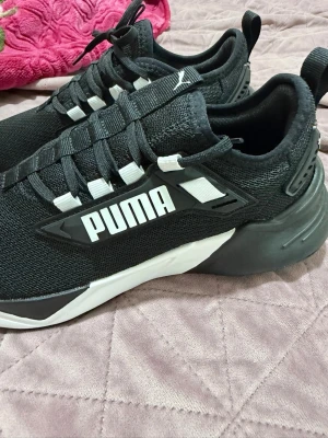 Svarta Puma sneakers med vit logga - Stilrena svarta sneakers från Puma med tydlig vit logga på sidan och vita detaljer vid snörningen. Ovandelen är i meshmaterial för bra andningsförmåga och sulan är i gummi med svart och vit design. Perfekta för en sportig och trendig look.