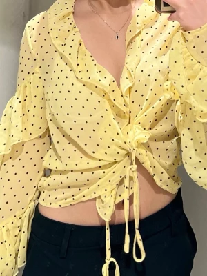 Polka dot blus med volanger - Söt gul blus med svarta prickar och volangkant längs urringningen. Blusen har långa ärmar och knytning framtill som ger en trendig look. Tillverkad i ett lätt och luftigt material som passar perfekt till vår och sommar.