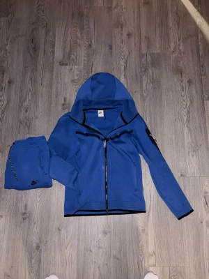 Blå Nike Tech - Snygga blå mjukisbyxor från Nike med matchande hoodie. Byxorna har resår i midjan och en dragkedjeförsedd ficka på sidan. Hoodien har hel dragkedja och svart kantband. Perfekt för chill eller träning, riktigt skön passform och stilren design.