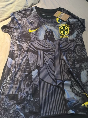 Nike Brasil fotbollströja svart/gul - Snygg svart fotbollströja från Nike med Brasil-logga och gult märke på bröstet. Framsidan har ett stort tryck av Kristusstatyn och detaljerade grafiska mönster i grått och svart. Korta ärmar och rund hals. Perfekt för dig som älskar fotboll och Brasilien.