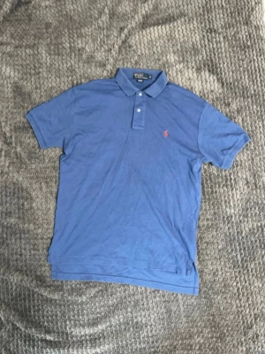 Blå pikétröja från Ralph Lauren - Klassisk blå pikétröja från Ralph Lauren med korta ärmar och krage. Tröjan har knäppning med två knappar och den ikoniska röda loggan broderad på bröstet. Perfekt för en avslappnad och stilren look. Lite längre i modellen. 