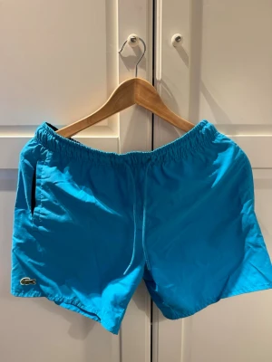 Blå badbyxor från Lacoste - Snygga blå badbyxor från Lacoste med klassisk krokodillogga på benet. Modellen har elastisk midja med snörning och sidofickor. Kommer med originalpåse från Lacoste. Perfekta för sommardagar vid stranden eller poolen.