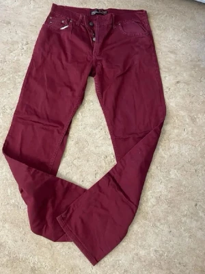 Vinröda bootcut jeans - Snygga vinröda jeans med bootcut passform och klassisk femficksdesign. Jeansen har knappgylf och är tillverkade i ett mjukt bomullstyg. Perfekta för dig som vill sticka ut med färg och stil.