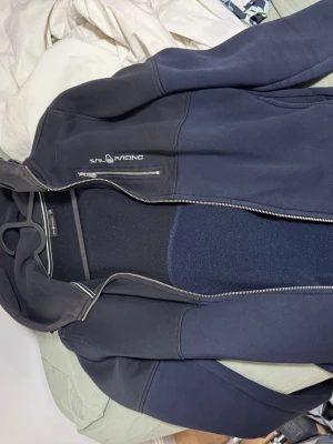 Mörkblå hoodie från Sail Racing - Mörkblå hoodie från Sail Racing med dragkedja och huva. Snygg logga på bröstet och praktiska fickor framtill. Tillverkad i mjukt material som känns skönt mot huden. Perfekt för en avslappnad och sportig stil.