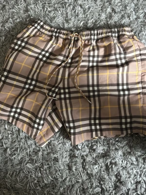Rutig shorts från Burberry med snörning - Säljer ett par klassiska rutiga shorts från Burberry i beige, brunt och gult. Shortsen har snörning i midjan och meshfoder på insidan. Perfekta för sommaren och har den ikoniska Burberry-looken. Skick: 9/10