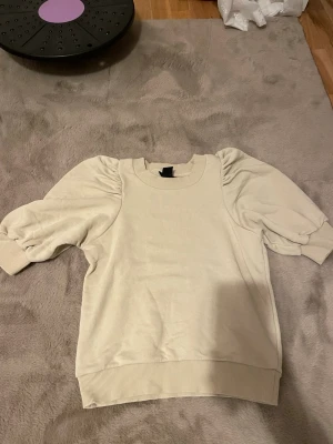 Beige topp med puffärmar - Trendig beige topp med puffiga kortärmar och breda muddar. Rund halsringning och avslappnad passform som ger en cool och modern look. Perfekt att styla med jeans eller kjol för en snygg outfit.