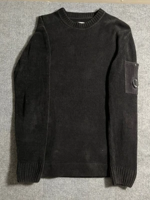 C.P. Company – Black Knit Lens Sweater (Size 48 / M–S) - Extremely rare C.P. Company knitted sweater in timeless black. Size 48 (fits like M/S), length 71 cm, width 50 cm – a perfect balance between comfort and tailored fit. Crafted from premium knit fabric, this piece delivers warmth, texture, and unmistakable luxury.  🖤 Condition: Excellent – no signs of wear or damage 💯 Authenticity: 100% genuine, easily verified on certilogo.com 📦 Shipping: Sent immediately after purchase – fast and reliable delivery!  This sweater stands out as one of the hardest