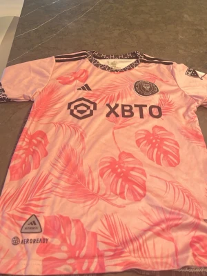 Inter Miami Messi 10  - Säljer en rosa Inter Miami matchtröja från Adidas med tropiskt bladmönster i olika rosa nyanser. Tröjan har svarta detaljer, klubbmärke på bröstet och sponsorloggor framtill. Tillverkad i lätt och ventilerande material med AEROREADY-teknologi.
