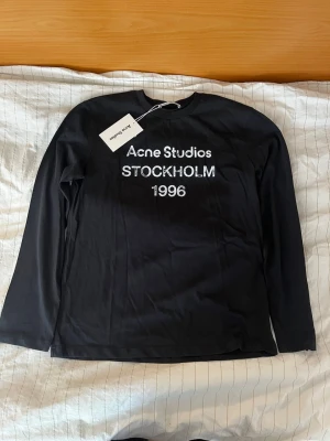 Svart långärmad topp Acne Studios - Svart långärmad topp från Acne Studios med tryck i vitt framtill där det står 'Acne Studios STOCKHOLM 1996'. Tillverkad i mjuk bomull och har en klassisk rund hals. Perfekt för dig som gillar minimalistisk och stilren design.