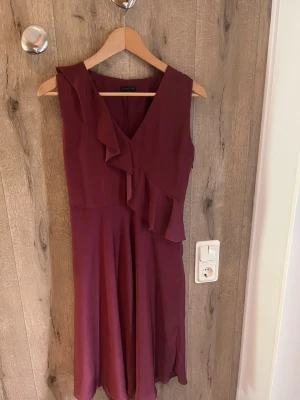 Skitsnygg Vinröd klänning  - Vinröd ärmlös klänning med omlott och volangdetalj framtill. Klänningen har v-ringning och är tillverkad i ett lätt, flytande tyg som ger ett elegant fall. Perfekt för dig som gillar stilrena och feminina plagg.