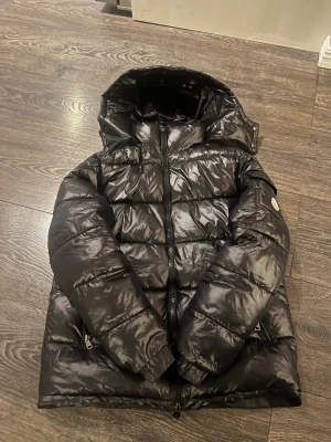Svart monc jacka - Snygg svart pufferjacka med glansig finish och huva. Jackan har dragkedja framtill, två sidofickor med dragkedja och quiltad design. Perfekt för kalla dagar när du vill hålla värmen och ändå se cool ut OBS en av jackfickorna är trasiga