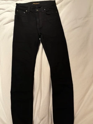 Svarta jeans från Nudie Jeans - Säljer ett par svarta jeans från Nudie Jeans med klassisk femficksdesign och raka ben. Jeansen har diskreta sömmar på bakfickorna och en liten logotyp på höger bakficka. Materialet är bomull med stretch för extra komfort.