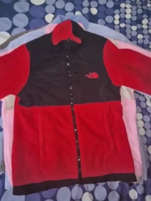 Röd och svart fleecejacka The North Face - Röd och svart fleecejacka från The North Face med hel dragkedja framtill och hög krage. Jackan har logga på bröstet och är perfekt för kyliga dagar. Klassisk passform och mjukt material.