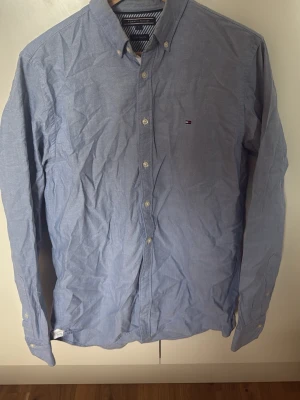 Blå skjorta från Tommy Hilfiger - Klassisk ljusblå skjorta från Tommy Hilfiger med button-down krage och diskret logga på bröstet. Skjortan har långa ärmar, vita knappar och är tillverkad i bomull. Perfekt för en clean och stilren look.