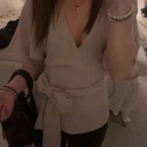 Beige omlottblus med knyt  - Säljer en stilren beige omlottblus från MW med knyt i midjan och korta, vida ärmar. Blusen har v-ringning och är elegant. Storlek 34. super snygg, skicka pris förslag skriv om du har några frågor eller funderingar