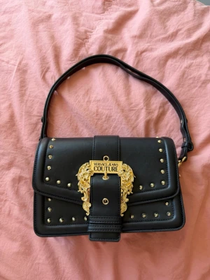 Svart Versace Jeans Couture handväska - Snygg svart handväska från Versace Jeans Couture med guldfärgade detaljer och nitar. Väskan har ett dekorativt spänne framtill, justerbart axelband och flera fack inuti, inklusive ett med dragkedja. Tillverkad i slätt konstläder för en lyxig känsla. Köpt på Zalando 2021. Köpt för 3800kr.