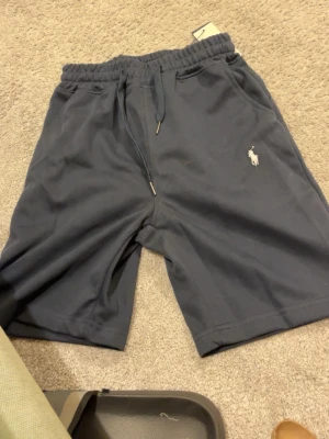 Mörkblå shorts  - Snygga mörkblå shorts med klassisk broderad logga på benet. Shortsen har elastisk midja med snörning och är tillverkade i mjukt material, perfekta för sommardagar. Två sidofickor för extra funktionalitet.