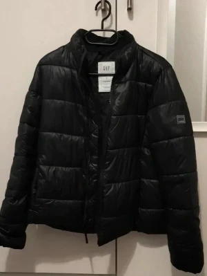 Svart pufferjacka från GAP - Snygg svart pufferjacka från GAP med quiltad design och hög krage. Jackan har dragkedja framtill och en diskret logotyp på ärmen. Perfekt för kalla vinterdagar och enkel att matcha med olika outfits.