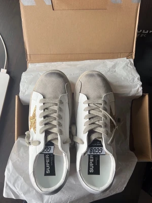 Golden Goose vita sneakers med glitterstjärna - Säljer ett par vita Golden Goose sneakers med beige mockadetaljer och en stor guldglittrig stjärna på sidan. Skorna har beige snören och sula samt GGDB STAR-tryck på sidan. Klassisk low-top modell i läder och mocka. Storlek 40 men känns mer som 39