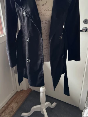 Svart trenchcoat med knytskärp - Stilren svart trenchcoat med dubbelknäppning och knytskärp i midjan. Klassisk krage och långa ärmar. Jackan har stora knappar framtill och är tillverkad i ett mjukt tyg som ger en snygg siluett.