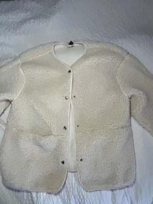 Krämvit teddyjacka med tryckknappar - Mysig krämvit teddyjacka i fluffigt material med rund halsringning och tryckknappar framtill. Jackan har en rak passform och är perfekt för lager-på-lager. Fodrad insida för extra komfort. Enkel och stilren design utan synliga fickor eller logga.