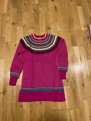 Mönstrad rosa stickad klänning stl S  - Säljer en långärmad stickad klänning från Dale of Norway i stark rosa med mönstrad ok i svart, vitt, gult och blått. Klänningen har ribbade muddar och dekorativa ränder vid ärmslut och nederkant. Perfekt för dig som gillar färg och nordisk design.