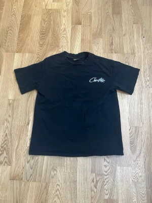 Svart Corteiz t-shirt med tryck - This black Corteiz t-shirt features a distinctive white reflective 'Cortiez' logo on the left chest and a larger reflective logo across the back, complemented by two reflective stripes on the shoulders. Crafted from comfortable cotton, its unique design makes it a standout streetwear piece. 
