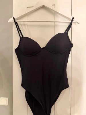 Svart body med bygel och smala axelband - Stilren svart body med vadderade kupor och bygel för extra stöd. Smala, justerbara axelband och en liten metallogo bak. Tillverkad i stretchigt material som sitter bekvämt och formar sig efter kroppen. Den är helt ny aldrig använd 