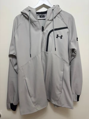 Grå hoodie från Under Armour - Snygg grå hoodie från Under Armour med dragkedja, svarta detaljer och logga på bröstet och ärmen. Huva med dragsko och två fickor framtill. Perfekt för en sportig och avslappnad stil.