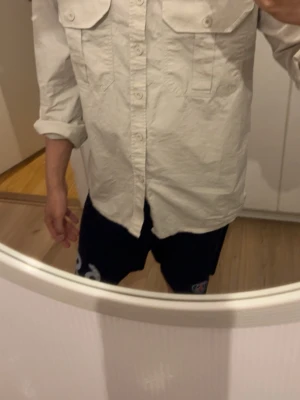 Beige skjorta med bröstfickor - Stilren beige skjorta med två stora bröstfickor och knappar framtill. Skjortan har lång ärm och klassisk krage, perfekt för en avslappnad men ändå snygg look. Tillverkad i mjuk bomull som känns skön mot huden.
