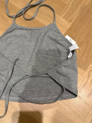 Grå omlottopp från NLY Trend - Snygg grå omlottopp från NLY Trend med tunna axelband och knyt i midjan. Toppen är ärmlös och har en croppad passform, perfekt för en trendig look. Tillverkad i mjukt bomullsmaterial för skön känsla.