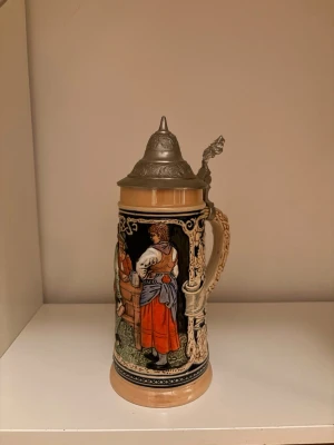 Tysk ölsejdel med lock och motiv - Säljer en klassisk tysk ölsejdel i keramik med detaljerat handtag och lock i metall. Sejdeln har färgglada motiv av personer i traditionella kläder och dekorerade mönster i beige, svart, rött och blått. Perfekt som samlarobjekt eller dekoration.
