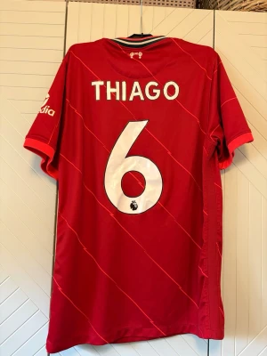 Liverpool matchtröja Thiago #6 - Röd Liverpool matchtröja från Nike med vita detaljer, tryck av 'Thiago' och nummer 6 på ryggen. Tröjan har klubbmärke, sponsorlogga och korta ärmar. Perfekt för fotbollsfans som vill stötta sitt lag.