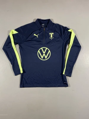 Malmö FF träningsset från Puma - Mörkblått träningsset från Puma med Malmö FF-logga och VW-tryck. Setet består av långärmad tröja med halv dragkedja och matchande byxor, båda med neongula detaljer längs ärmar och ben. Tillverkat i funktionsmaterial som passar perfekt till fotbollsträning.
