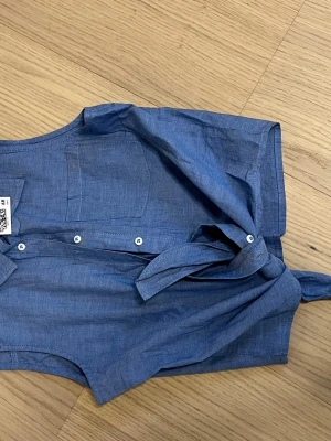 Blå söt kortärmad knytblus med knappar  - ny! - Snygg blå blus i mjuk bomull. Blusen har korta ärmar, rund halsringning och knäppning med vita knappar fram. Det finns även ett knytband i midjan. Ny!  (ord. pris 99 sek, säljer för 4944