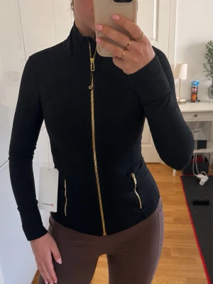 Svart jacka med guldiga dragkedjor lululemon define jacket 2 S svart guld gold black  - Snygg svart jacka lululemon med figurnära passform och guldiga dragkedjor framtill och vid fickorna. Jackan har hög krage och långa ärmar, perfekt för en stilren look. Passar till många olika outfits och är lätt att matcha. Stl 2 passar S 