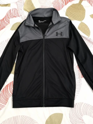 Svart träningsjacka från Under Armour - Snygg svart träningsjacka från Under Armour med grå panel upptill och dragkedja hela vägen. Jackan har hög krage, långa ärmar och är tillverkad i ett lätt och stretchigt material som passar perfekt till träning eller uppvärmning.