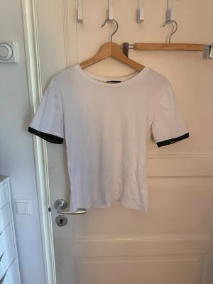 Vit t-shirt med glittriga ärmslut - Vit t-shirt med rund hals och korta ärmar. Ärmsluten har en snygg svart glittrig kant som ger en cool kontrast till den enkla designen. Perfekt för dig som vill ha en basic t-shirt med en twist.