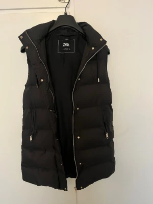 Svart dunväst från Zara med huva - Stilren svart dunväst från Zara med huva och dragkedja framtill. Västen har två fickor med dragkedja och quiltad design som ger en puffig look. Perfekt för lager-på-lager och passar till många outfits. Materialet är syntet med dunliknande fyllning.