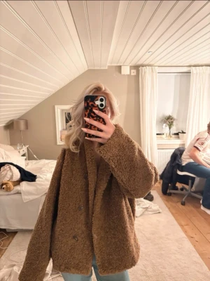 Brun oversized teddyjacka från Gina Tricot  - Mysig och oversized teddyjacka i brun färg med dubbelknäppning framtill. Jackan har en fluffig och mjuk textur som ger en trendig look. Perfekt för att hålla värmen under kyliga dagar. Passar dig som gillar en avslappnad och cool stil.