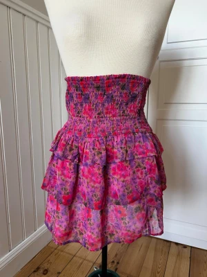 Blommig volangkjol från BikBok - Supersöt kort kjol från BikBok i blommigt mönster med rosa och lila toner. Kjolen har smock i midjan och dubbla volanger för en extra flowy känsla. Perfekt för dig som gillar färg och vill sticka ut lite extra.