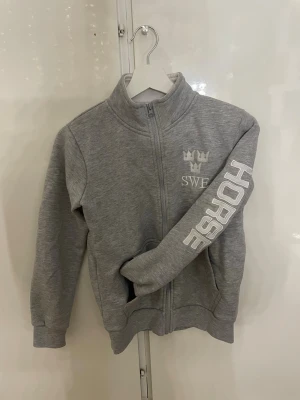 Grå rid zip-hoodie med tryck SWE HORSE - Grå rid zip-hoodie med hög krage och dragkedja framtill. På bröstet finns ett vitt tryck med tre kronor och texten SWE, samt stort HORSE-tryck längs ena ärmen. Mjuk bomullsmix och ribbade muddar. Perfekt för dig som gillar hästar och sportig stil.