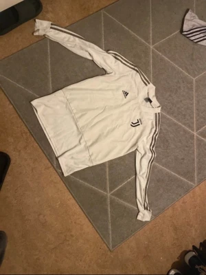Vit Juventus träningströja Adidas - Vit långärmad träningströja från Adidas med Juventus-logga på bröstet och klassiska tre ränder längs ärmarna. Perfekt för fotbollsträning eller chill. Tröjan har rund hals och är gjord i ett lätt och bekvämt material.