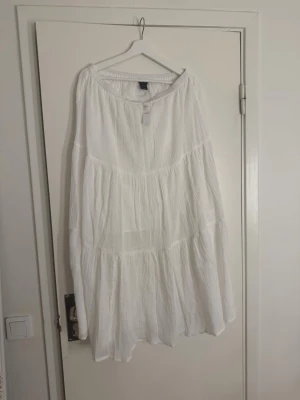 Vit volangkjol i bomull från H&M - Lång, luftig kjol i vit bomull från H&M med flera volanglager. Kjolen har en avslappnad och flowy passform, perfekt för sommar och vår. Resår i midjan för extra komfort och enkel styling.