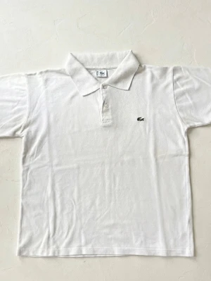 Vit pikétröja från Lacoste - Klassisk vit pikétröja från Lacoste med korta ärmar och krage. Tröjan har knappar vid halsen och den ikoniska lilla krokodilloggan på bröstet. Tillverkad i mjuk bomull som känns skön mot huden.