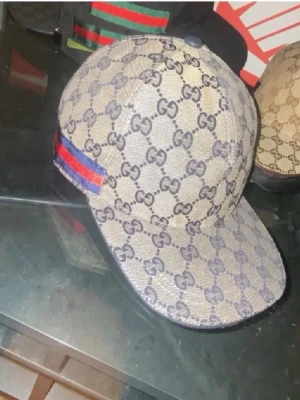 Gucci keps med GG-mönster och röd-blå rand - Snygg keps från Gucci med klassiskt GG-monogram i beige och blått. Kepsen har en böjd skärm och en ikonisk röd-blå rand på sidan. Tillverkad i canvasmaterial med justerbar rem baktill. Perfekt för dig som vill sticka ut med en lyxig accessoar.