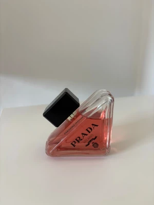 Prada Paradoxe Eau de Parfum - Unik trekantig parfymflaska från Prada med rosa doftvätska och svart fyrkantigt lock. Flaskan är tillverkad i glas och har en modern, stilren design med tydlig Prada-logga på framsidan. Perfekt för dig som gillar lyxiga och trendiga dofter.